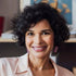 Smiling woman therapist with curly dark hair wearing a light pink shirt and purple earrings in a cozy indoor setting